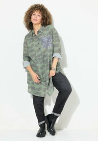 Groene oversized shirt met een patroon en een pailletten zak, gecombineerd met donkere jeans en zwarte enkellaarsjes. Heeft opgerolde mouwen en een gebogen zoom.