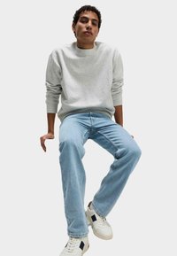Sudadera gris, jeans azul claro y zapatillas blancas con acentos negros. Corte relajado, puños acanalados y diseño sencillo. Posición sentada.