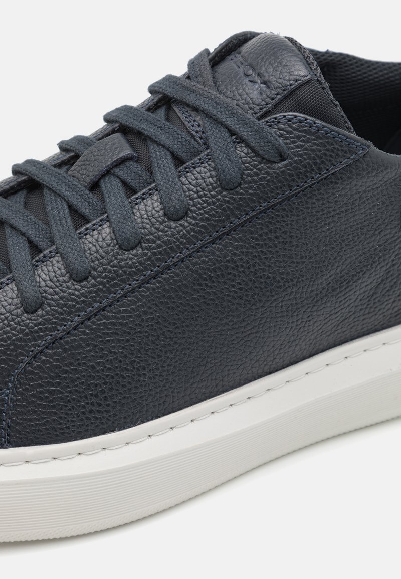 Baskets en cuir bleu foncé avec surface texturée, semelle en caoutchouc blanche, et lacets plats, présentant des coutures discrètes le long des bords.