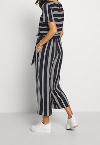 Mujer vestida con un mono azul marino con rayas verticales y horizontales blancas, mangas cortas, bolsillos laterales y zapatillas de plataforma blancas, posando de lado.