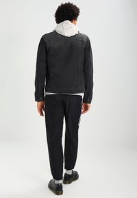 Svart denimjacka med en avslappnad passform, grå huvtröja under, och svarta träningsbyxor med vita sidostråk. Svarta skor fulländar looken.