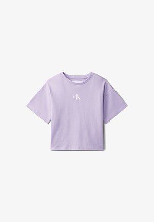 Jong meisje staat met hand in haar zak, draagt een lichtpaarse T-shirt met korte mouwen, een licht denim rokje en zwarte slippers, tegen een grijze achtergrond.