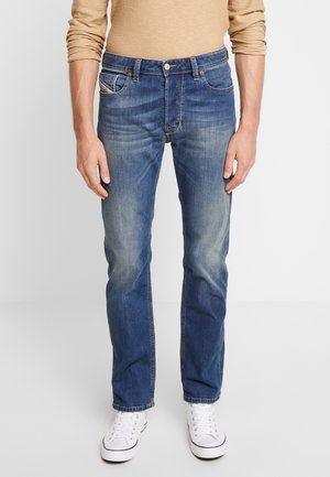 Mand iført blå falmede jeans, beige langærmet skjorte og hvide sneakers stående mod en ensfarvet lys baggrund.
