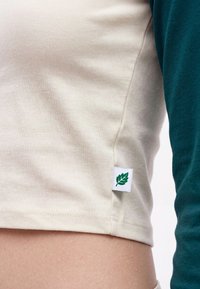 Gros plan d'un haut en tissu côtelé crème avec une manche longue verte et une petite étiquette affichant un logo de feuille verte près de l'ourlet.