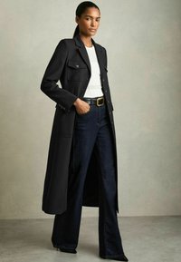 Palton lung negru, cu buzunare frontale, purtat peste un top alb și pantaloni din denim închiși, evazați. Are un design clasic și o croială ajustată.
