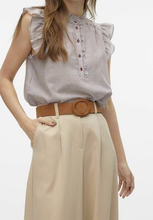 Vrouw draagt een mouwloze beige gestreepte blouse met ruches, gestopt in een beige broek met hoge taille en een brede bruine riem.