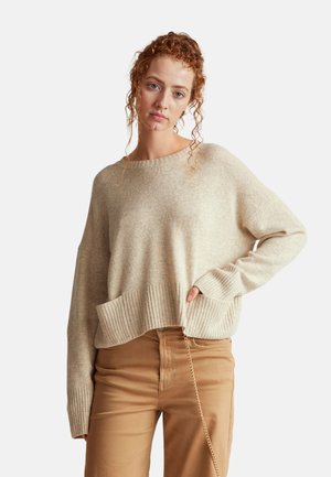 Suéter de punto beige con dobladillo y puños acanalados, que presenta un corte holgado y un escote amplio, combinado con pantalones marrones y un detalle de cadena.