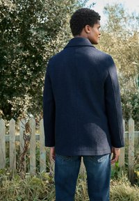 Marineblauer Wollmantel mit Reverskragen, geradem Schnitt und minimalen Nähten, kombiniert mit dunklen Jeans in einem Outdoor-Setting.