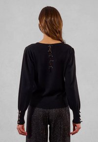 Donna di spalle che indossa una maglia nera a maniche lunghe con nastrini neri legati sul retro e sui polsini delle maniche.