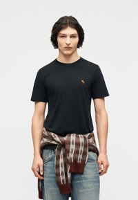 Νεαρός άνδρας με μαύρο κοντομάνικο πουκάμισο, μικρός κεντημένος αλκης στο στήθος, καρό καφέ και λευκό πουλόβερ δεμένο στη μέση, ανοιχτόχρωμο μπλε τζιν.