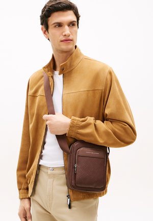 Tânăr purtând o jachetă bej și pantaloni bej, ținând o geantă crossbody maro Tommy Hilfiger, cu o curea peste umăr.