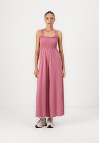 Rosa ärmelloser Jumpsuit mit einem gerafften Oberteil und weiten Beinen, aus weichem Stoff gefertigt. Trägt dazu helle Sneakers vor einem schlichten Hintergrund.