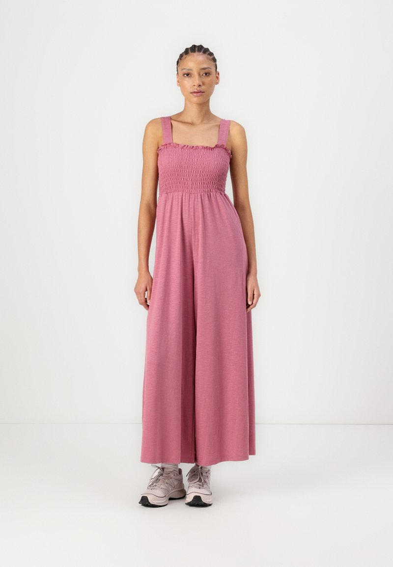 Rosa ärmelloser Jumpsuit mit einem gerafften Oberteil und weiten Beinen, aus weichem Stoff gefertigt. Trägt dazu helle Sneakers vor einem schlichten Hintergrund.