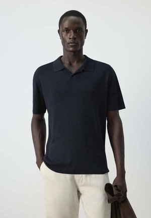 Hombre vestido con una camiseta polo azul marino de manga corta y pantalones beige claros, sosteniendo una chaqueta marrón en su mano izquierda sobre un fondo liso.