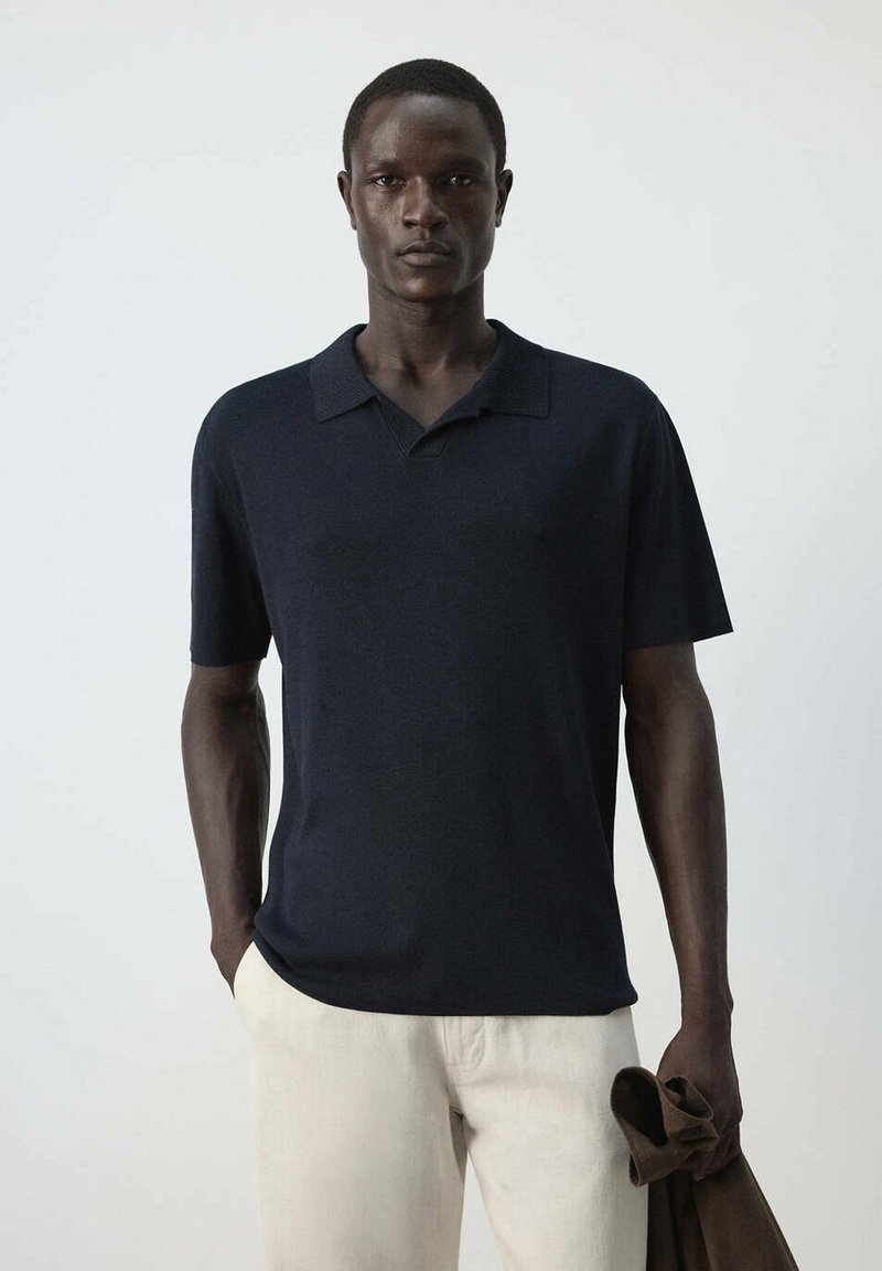 Homme portant un polo bleu marine à manches courtes et un pantalon beige clair, tenant une veste marron dans sa main gauche devant un fond uni.