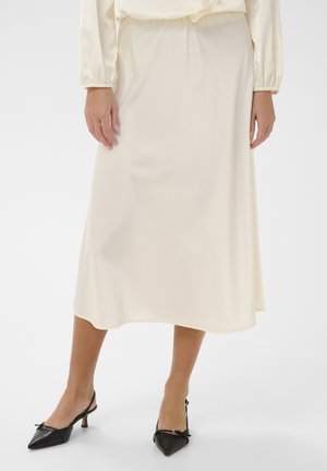 Frau trägt einen cremefarbenen Satin-Midirock und eine passende langärmelige Bluse mit schwarzen Slingback-Pumps mit Spitze und Schleifen.