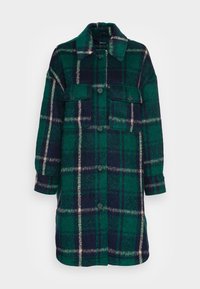 Grön och marinblå rutiga långkappa i texturerad tyg. Har en krage, framknäppning och två bröstfickor med klaffstängning.