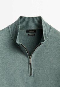 Pull-over en maille verte avec un col haut côtelé et une fermeture éclair métallique à demi-curseur, affichant l'étiquette de la marque et l'étiquette "Fabriqué en Chine" à l'intérieur du col.