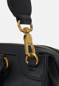 Dettaglio della tracolla di una borsa in pelle nera che presenta un clips e un anello in metallo dorato, con cuciture visibili e una cerniera lungo il bordo.