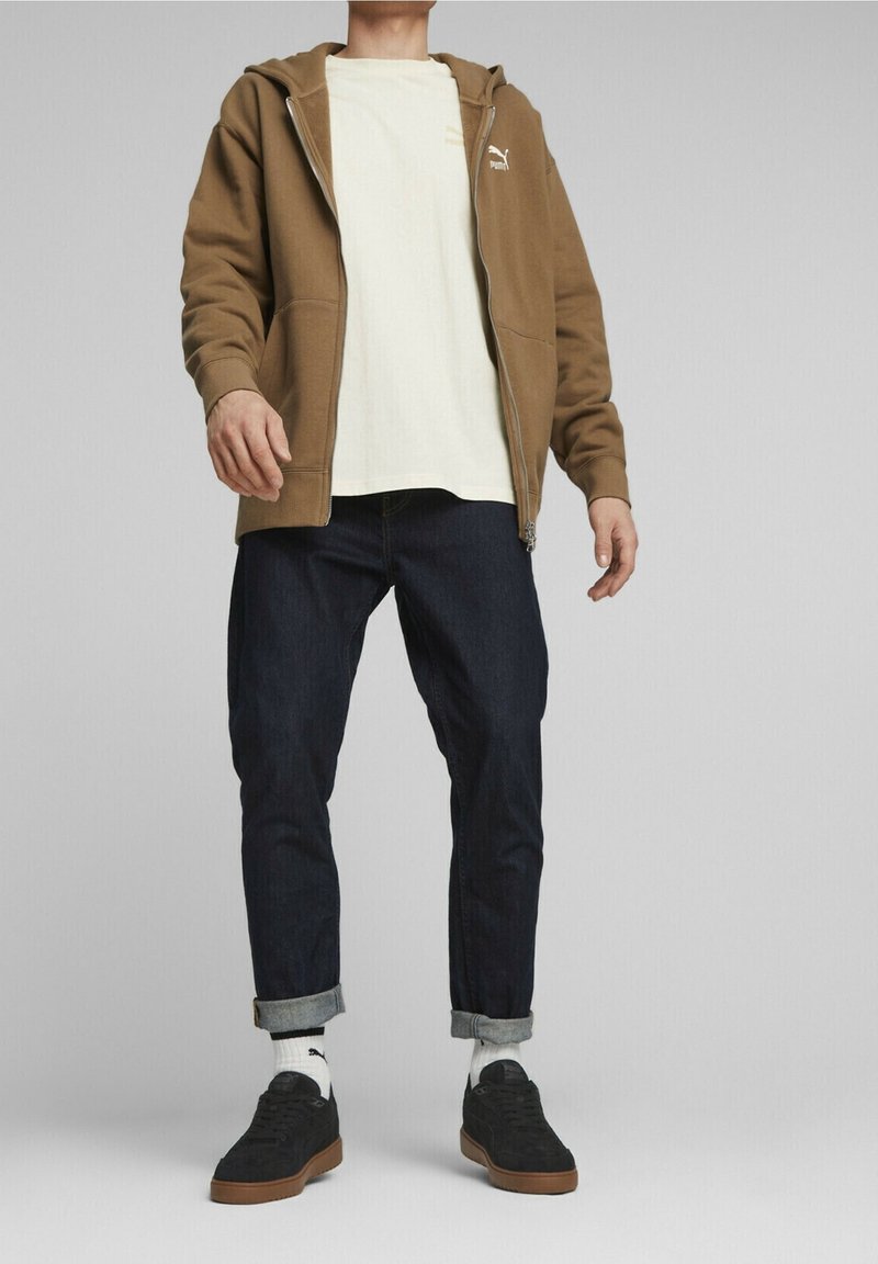 Brun zip-up hoodie över en ljusfärgad t-shirt, tillsammans med mörka jeans och svarta sneakers med beige sulor och vita strumpor.