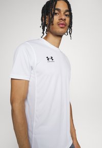 Īsām piedurkņu balta sporta krekls, izgatavots no gludas, elpojošas auduma, ar melnu Under Armour logotipu kreisajā krūtīs.