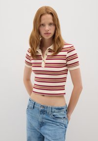 Camisa polo de manga corta a rayas en crema, rojo y naranja, con un diseño ajustado, textura acanalada y una tapeta de dos botones. Combinada con vaqueros azules.