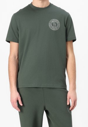 Homme portant une chemise à manches courtes vert foncé et un pantalon assorti avec un logo rond Lacoste Paris sur la poitrine et la cuisse.