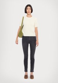 Gewebtes cremefarbenes Strickoberteil, schwarze schmale Jeans und braune Mokassins. Das Modell hält eine olivgrüne Wildledertasche; minimalistisches Design mit gerippter Textur.