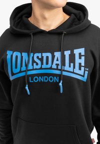 Zwarte hoodie met blauwe tekst "LONSDALE LONDON". Voorzien van een koord met blauwe punten en een voorzak. Gemaakt van zachte stof.