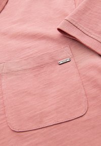 Světle růžový oděv s krátkým rukávem, který se pyšní jemnou texturou a přední kapsou se stříbrnou značkovou tagem. Detaily šití jsou decentní.