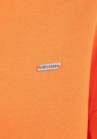 T-shirt orange avec une étiquette de marque rectangulaire grise portant l'inscription "SMILODOX" en noir. Le tissu présente une texture lisse et uniforme.