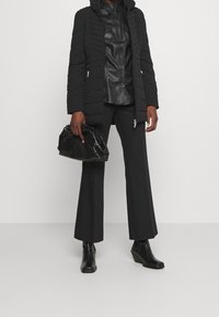 Cappotto imbottito nero con cappuccio, abbinato a una camicia in pelle nera con bottoni, pantaloni neri svasati e stivali neri alla caviglia. Borsa dalla texture lucida.