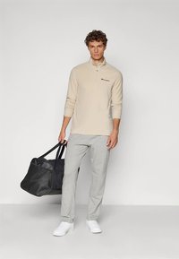 Mann i beige zip-up genser, grå joggebukser og hvite sneakers, holder en stor svart duffelbag ved sin side.
