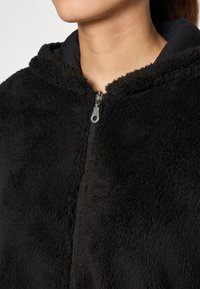 Felpa con zip nera in pile, con un'esterno testurizzato, dotata di una fodera interna nera e una zip argentata. Dettaglio del cappuccio incapsulato visibile.