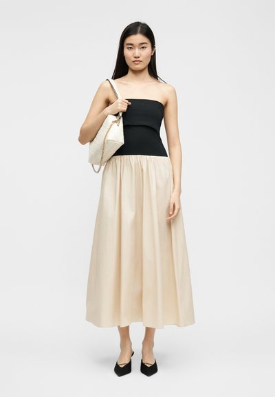 Γυναίκα που φοράει μαύρο τοπ χωρίς τιράντες και μπεζ φούστα έως τον αστράγαλο, κρατώντας λευκή τσάντα στον ώμο, στέκεται μπροστά σε απλό φόντο.