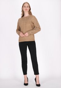 Beige kábeles kötésű pulóver hosszú ujjakkal és elülső zsebbel, fekete slim-fit nadrággal és fekete magassarkú cipővel párosítva.
