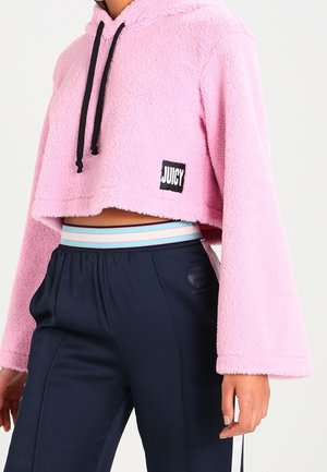 Sweat à capuche en polaire rose raccourci avec cordons noirs et écusson "JUICY", associé à un pantalon bleu marine avec une ceinture rayée pastel.