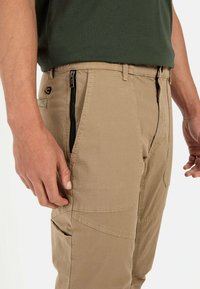 Beige Cargo-Hosen mit glatter Textur, ausgestattet mit seitlichen Reißverschlüssen, aufgesetzten Taschen und verstärkten Nähten. Kombiniert mit einem grünen Hemd.