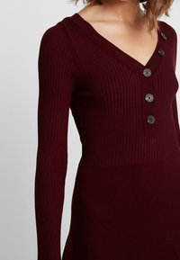 Abito a coste bordeaux con maniche lunghe, scollatura a V e cinque bottoni decorativi sul davanti per una silhouette aderente.