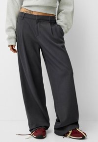 Pantaloni grigi a gamba larga con un design sartoriale, caratterizzati da pinces frontali, tasche laterali e una texture liscia, abbinati a sneaker rosse.