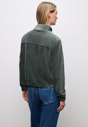 Giacca di velluto verde scuro con colletto alto, polsini elasticizzati e design cropped, abbinata a jeans in denim blu, vista da dietro.