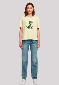 Gelbes Baumwoll-T-Shirt mit kurzen Ärmeln und einem grünen Dinosaurier-Print auf der Vorderseite, kombiniert mit blauen Jeans aus Denim und weißen Turnschuhen.