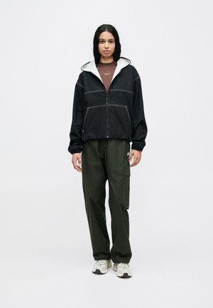 Giacca nera con zip e cappuccio grigio, con cuciture bianche; abbinata a pantaloni cargo verde oliva e sneaker beige.