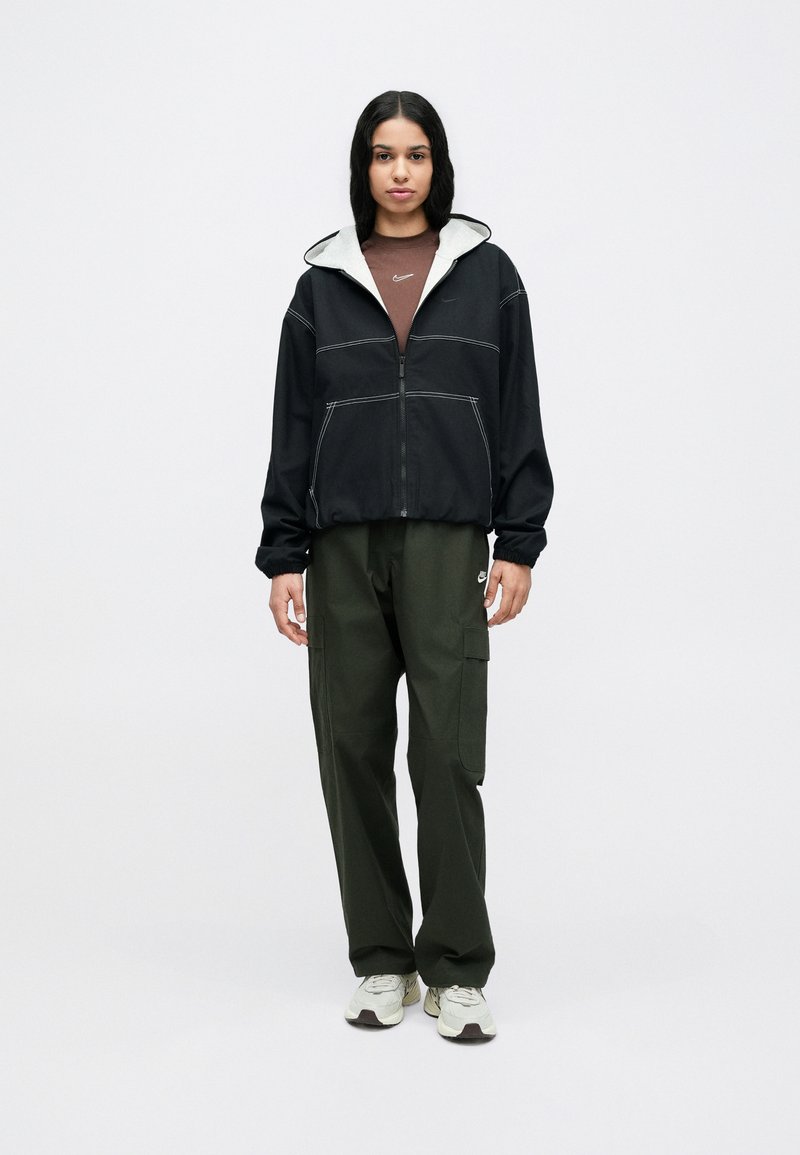 Μαύρο μπουφάν με φερμουάρ και γκρι κουκούλα, με λευκή ραφή· σετ με χακί παντελόνι cargo και μπεζ αθλητικά παπούτσια.