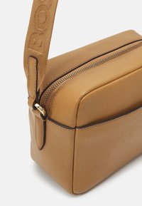 Hnědá kožená crossbody taška obdélníkového tvaru, s uzavíratelným zipem a popruhem z tkaniny s logem značky. Texturovaný povrch s jemným švem.