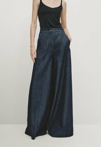 Femme portant un débardeur noir et un pantalon en denim bleu foncé, taille haute et à jambes larges, les mains dans les poches.