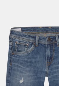 Jeans en denim avec un délavage bleu, présentant un léger effet usé, un design à cinq poches, des rivets en cuivre et des accents brodés à la taille.
