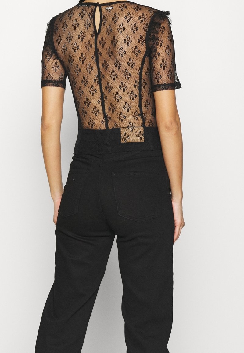 Haut en dentelle noire à manches courtes, présentant un motif floral et un détail dos avec ouverture en forme de goutte, associé à un jean noir taille haute.