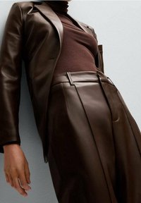 Osoba ubrana w brązową skórzaną marynarkę i dopasowane skórzane spodnie wysokiej talii oraz brązowy sweter z golfem, na jasnym tle.