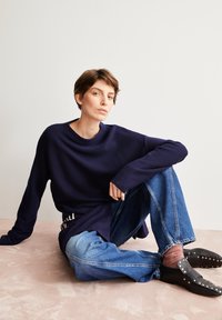 Marineblauer Langarmpullover, lockere Passform, kombiniert mit hellblauen weiten Jeans. Schwarze Schuhe mit silbernen Nieten und strukturierten Socken.